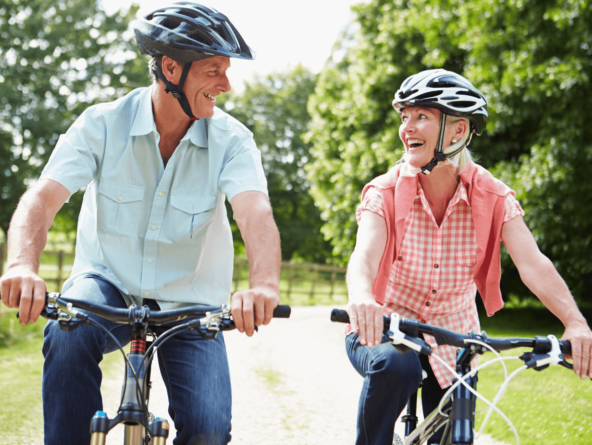 A smiling older couple riding bicycles on a sunlit park path, each wearing a black bicycle helmet. The man wears a light blue short-sleeve button-up shirt and jeans, while the woman wears a coral pink sleeveless shirt over a red-and-white gingham top with dark jeans.