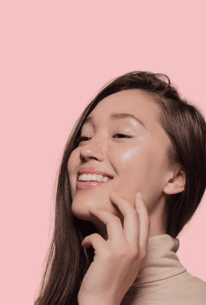 Woman smiling and touching her cheek, showing clear glowing skin, against a soft pink background.